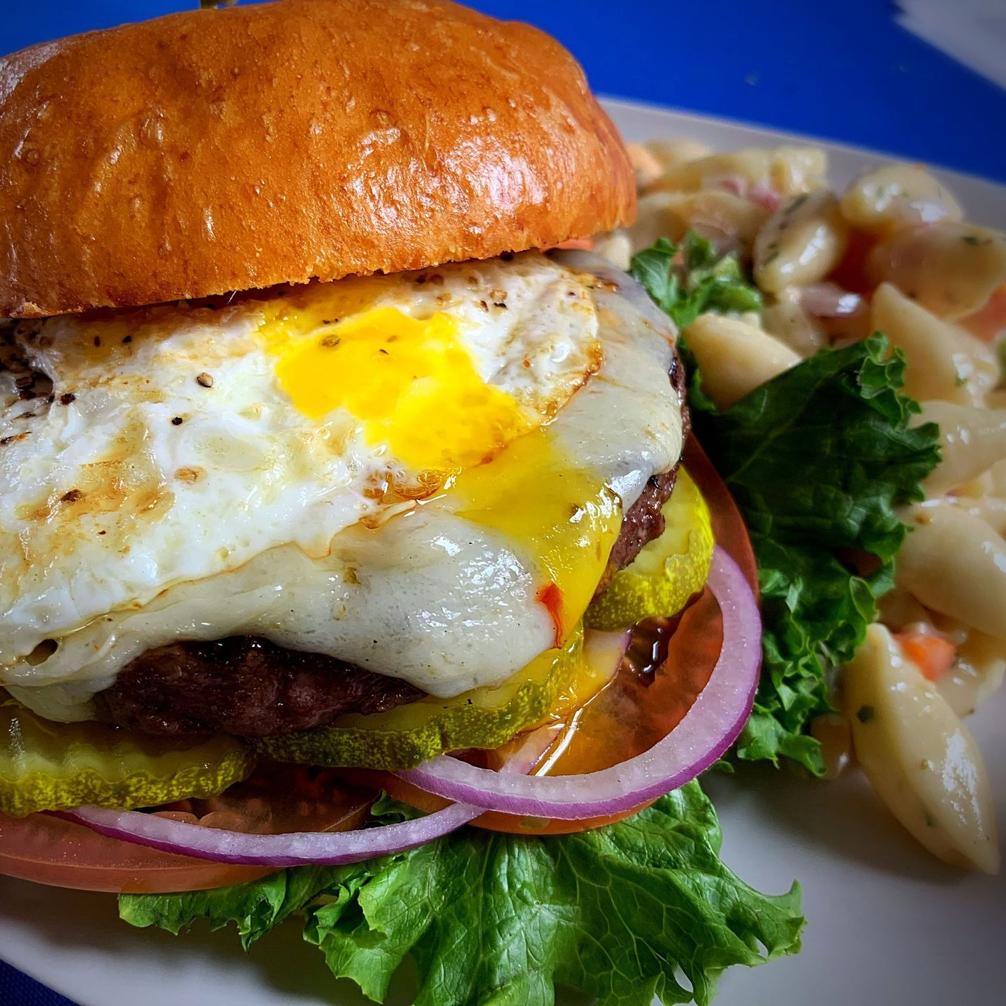 Burger with egg, pickles, and red onion on a bun, served with pasta salad.