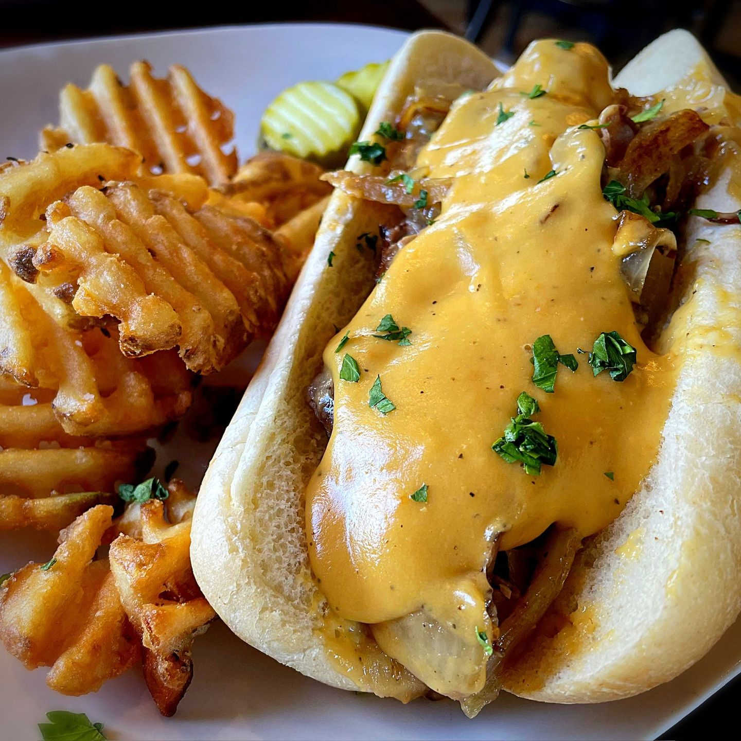 Philly sandwich with cheese sauce, waffle fries, and pickles on a white plate.
