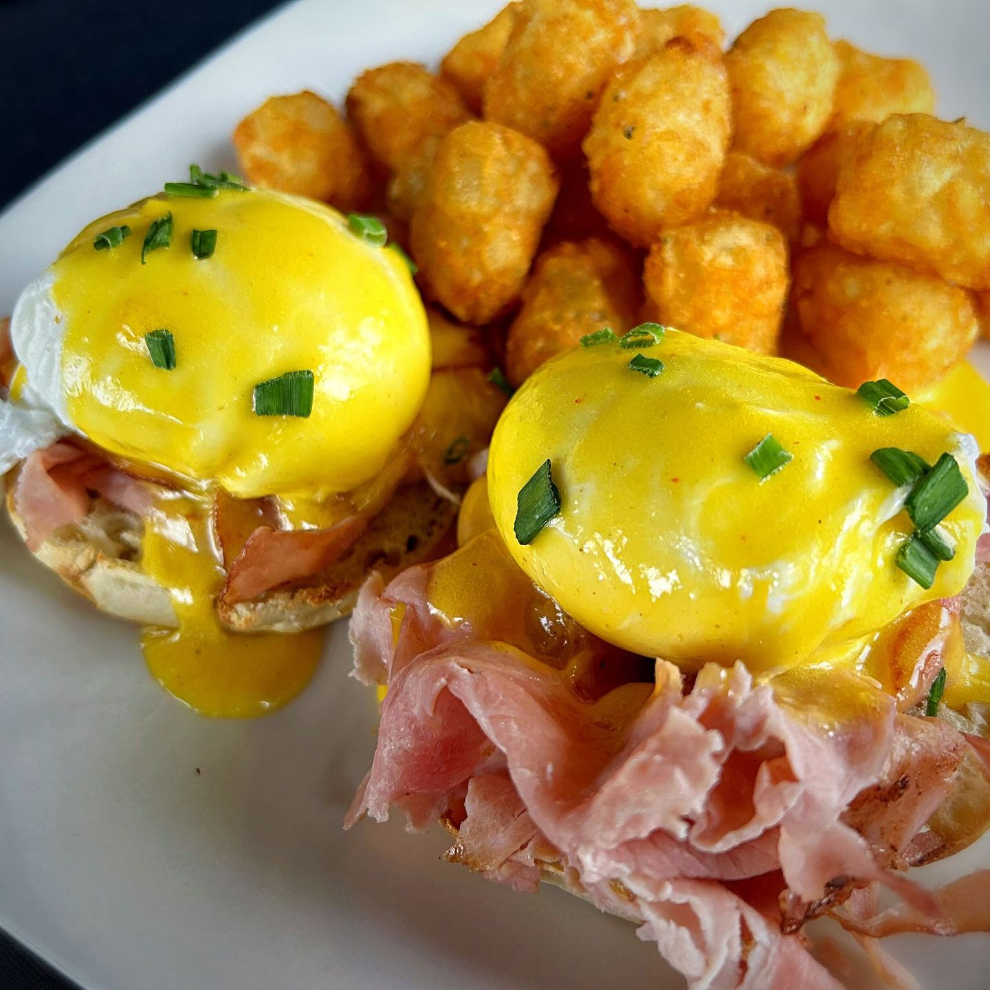 Eggs Benedict with tater tots on a white plate. Yellow hollandaise sauce over poached eggs, ham, and English muffins.