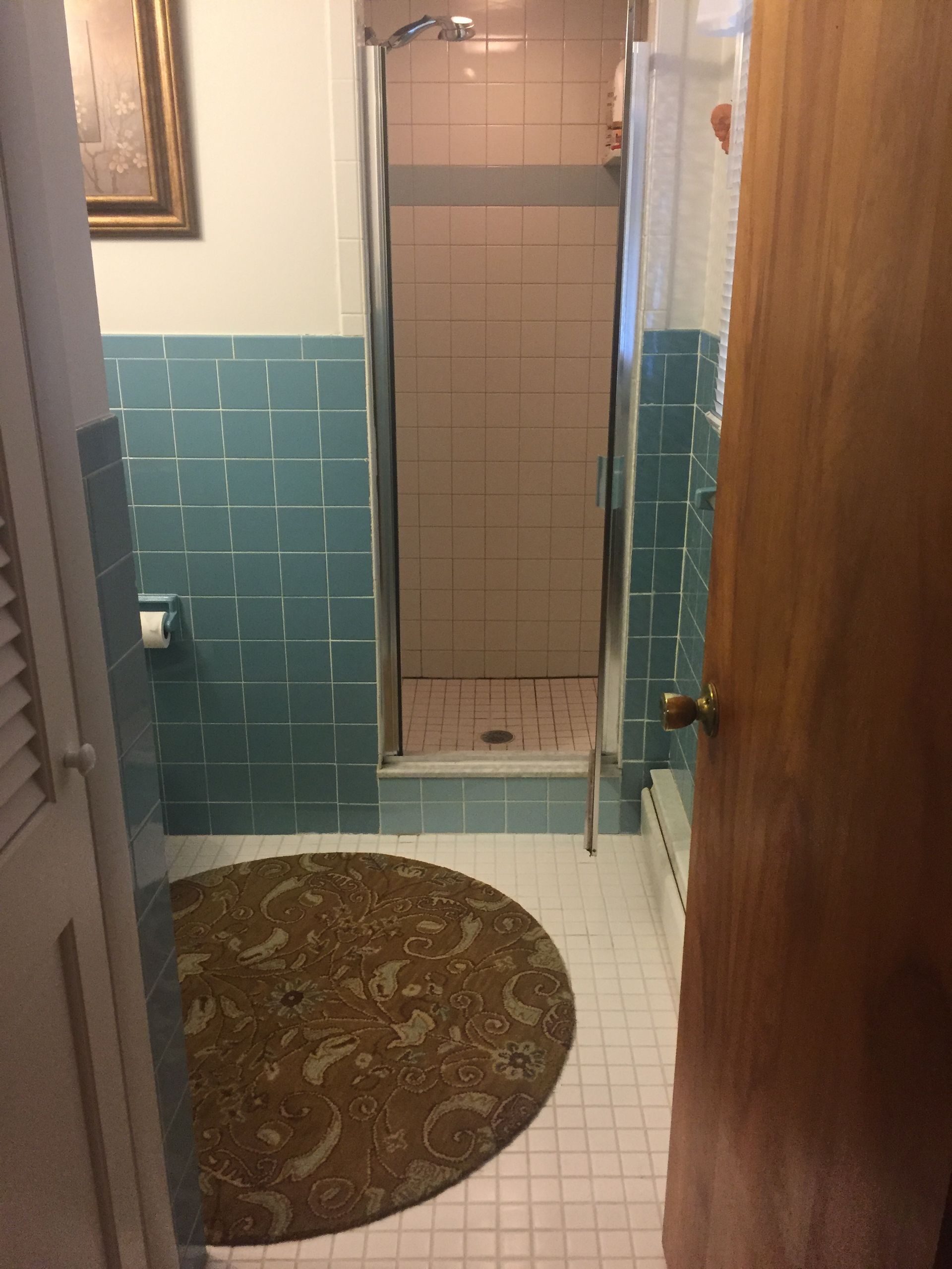Small bathroom with blue tile walls, white floor, shower stall, and round rug; door open.