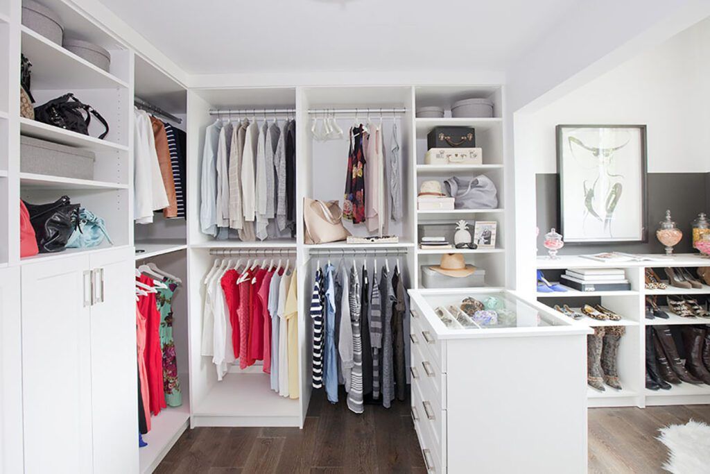 Bright walk-in closet with white shelving, hanging clothes, and shoes organized around a central island.