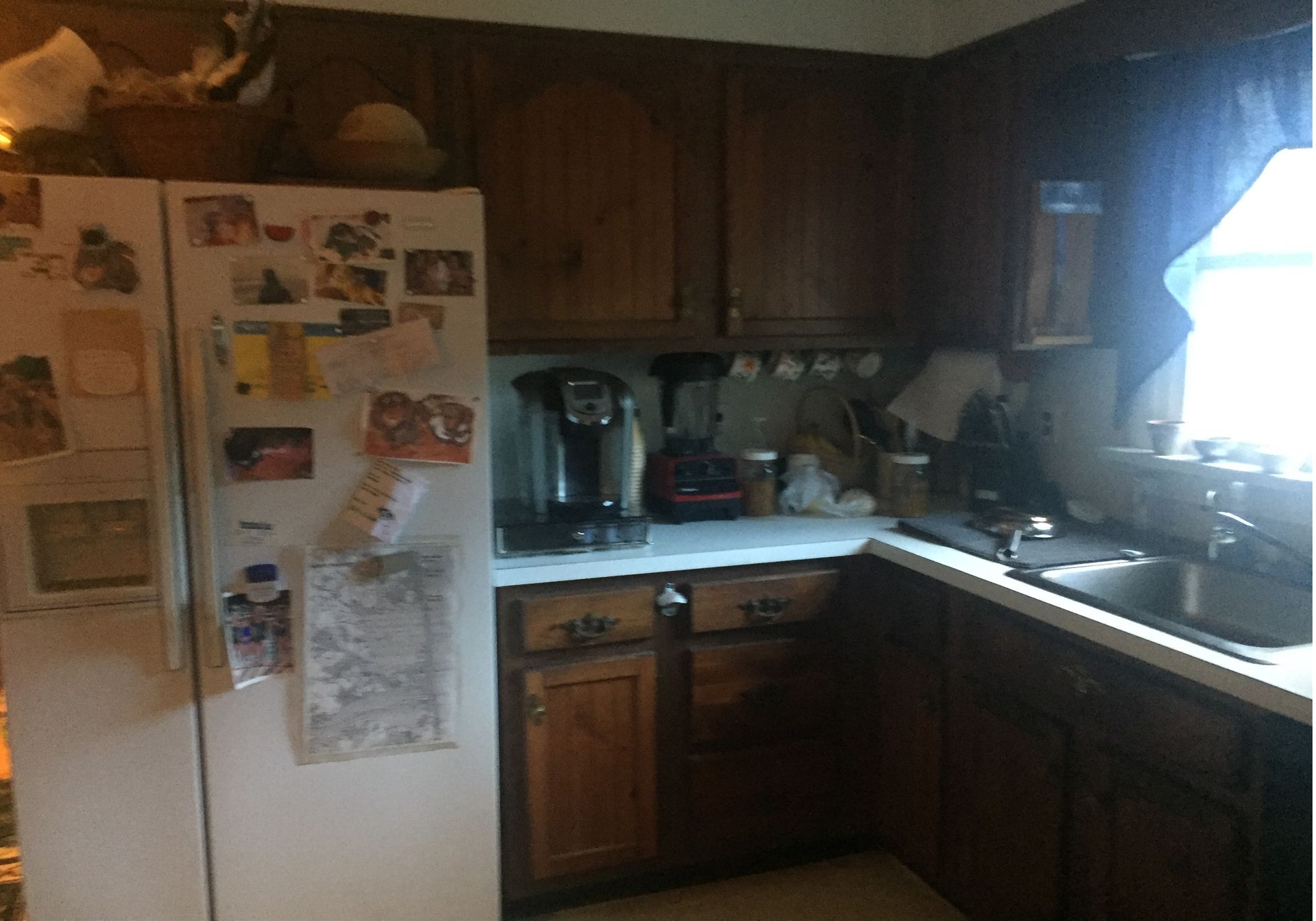 Dim kitchen with wooden cabinets, white fridge covered in notes, and countertop appliances by a sunlit sink.