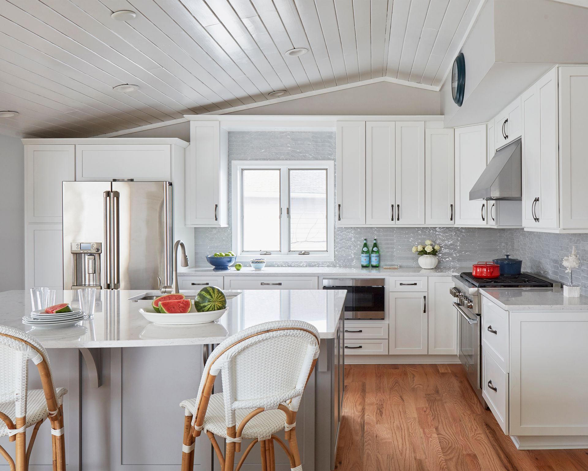 Bright white kitchen with island, stainless fridge, and wicker chairs by a sunlit window.