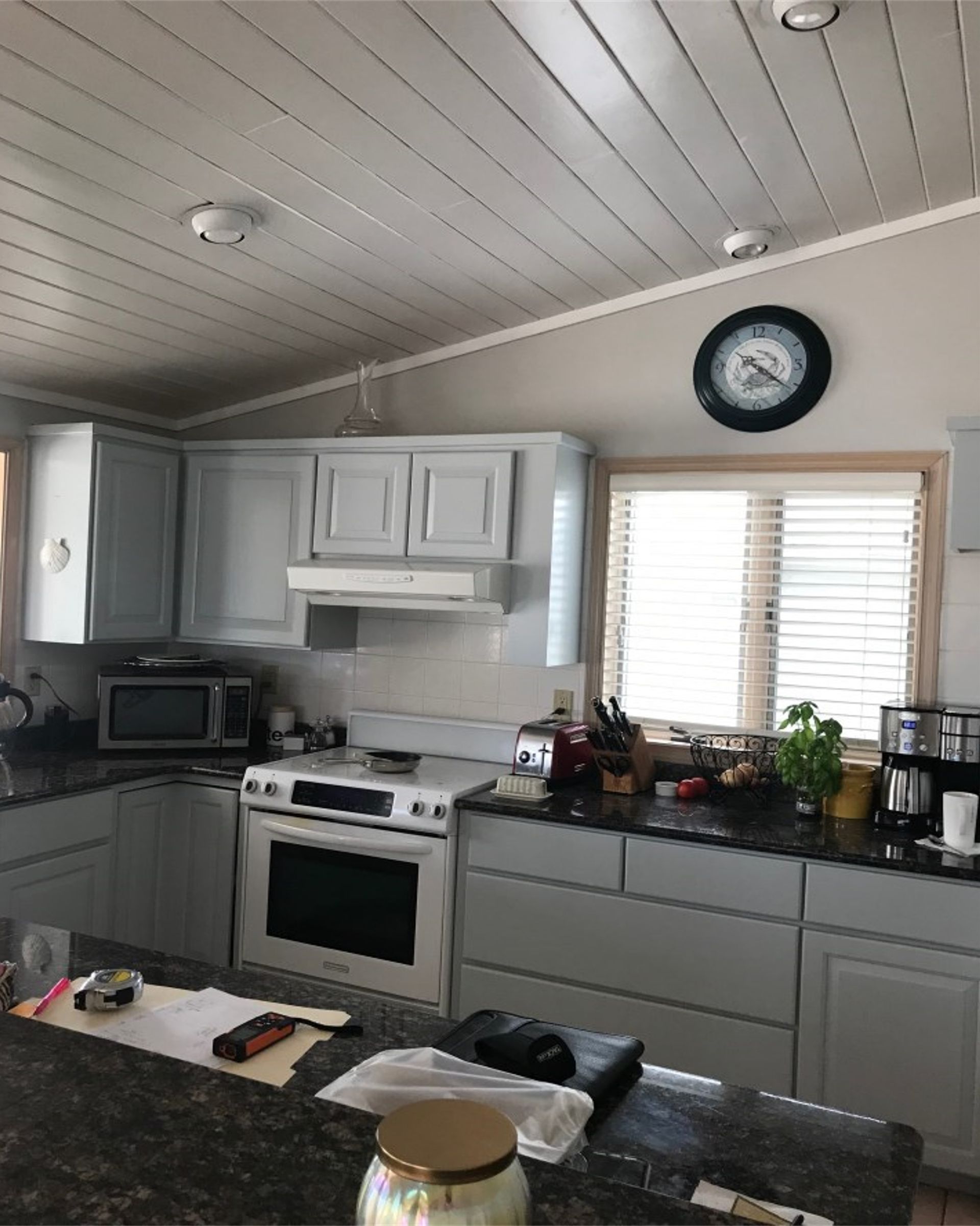 Bright kitchen with white cabinets, black countertop, stove, microwave, and a wall clock above the window.