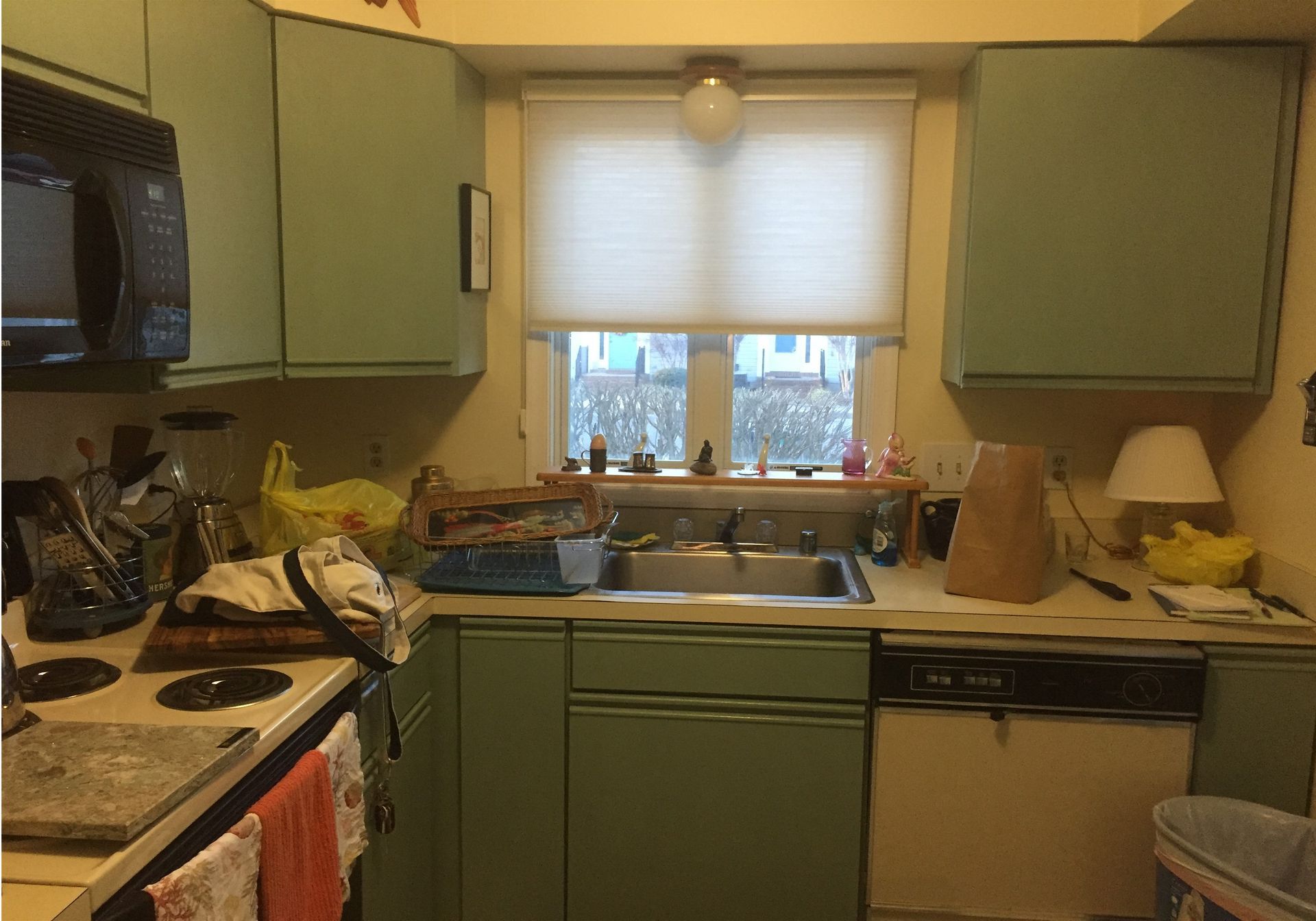 Small kitchen with green cabinets, cluttered counters, sink under window, and stove on the left.
