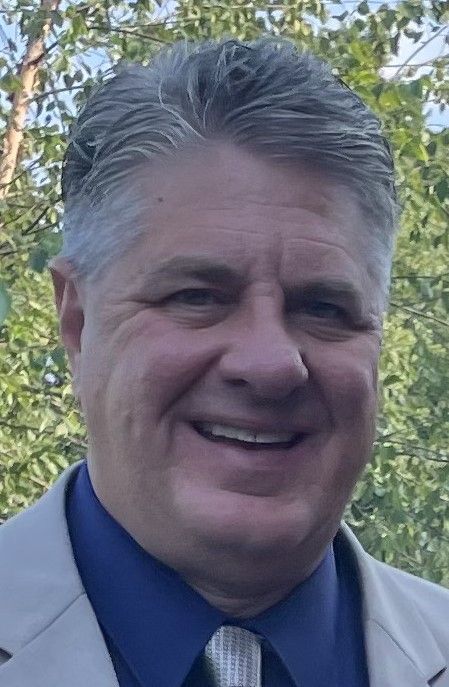 Smiling man in a beige jacket and blue shirt outdoors with trees in the background