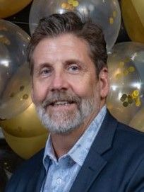 Smiling man in a blue blazer and light shirt in front of gold and silver balloons