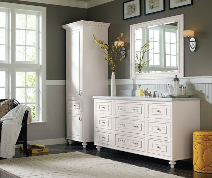 Bright white bedroom with dresser, armoire, large mirror, and yellow ottoman beside tall windows.