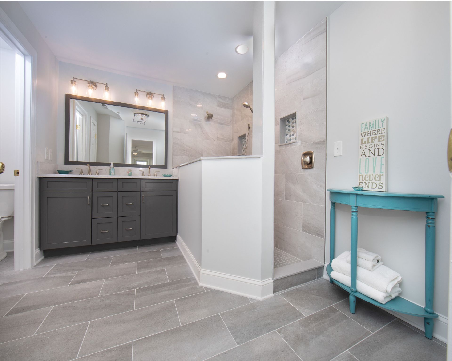 Bright gray bathroom with double vanity, large mirror, tiled shower, and turquoise side table.