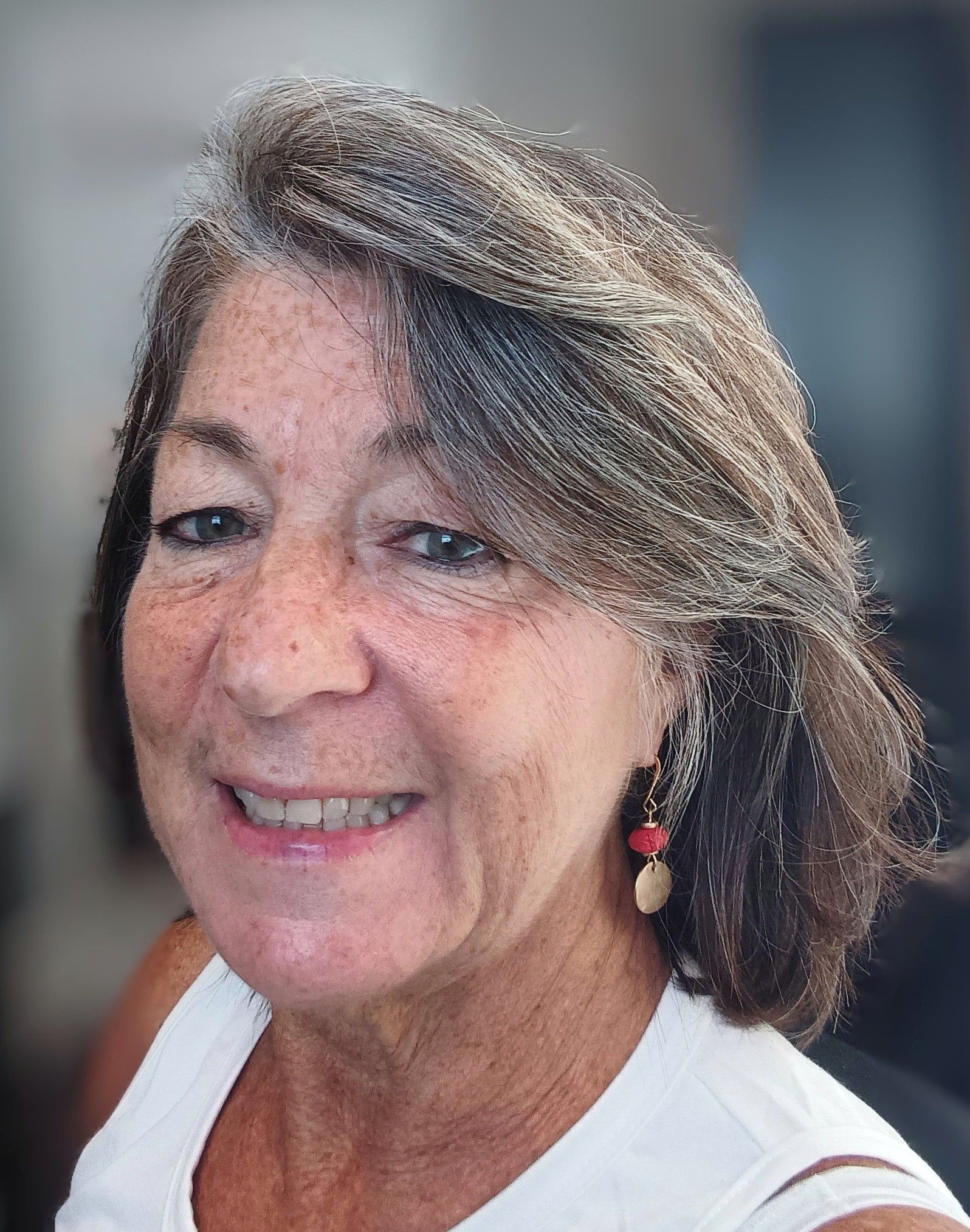 Smiling woman with shoulder-length brown hair wearing a white top and red earrings against a blurred indoor background