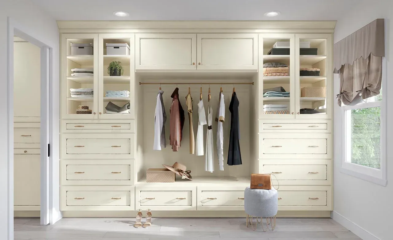 Built-in cream closet with hanging clothes, drawers, shelves, and a bench in a bright bedroom
