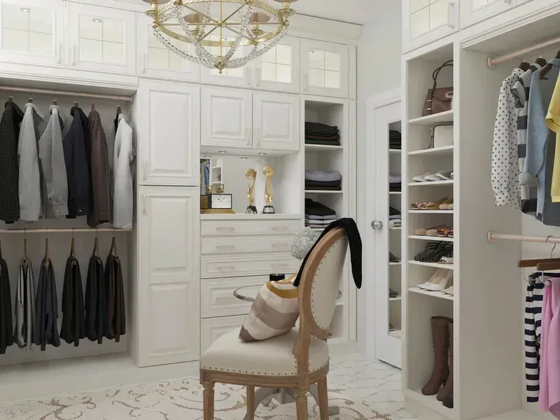 Bright white walk-in closet with hanging clothes, shelves, and a chair with a hat and bag.
