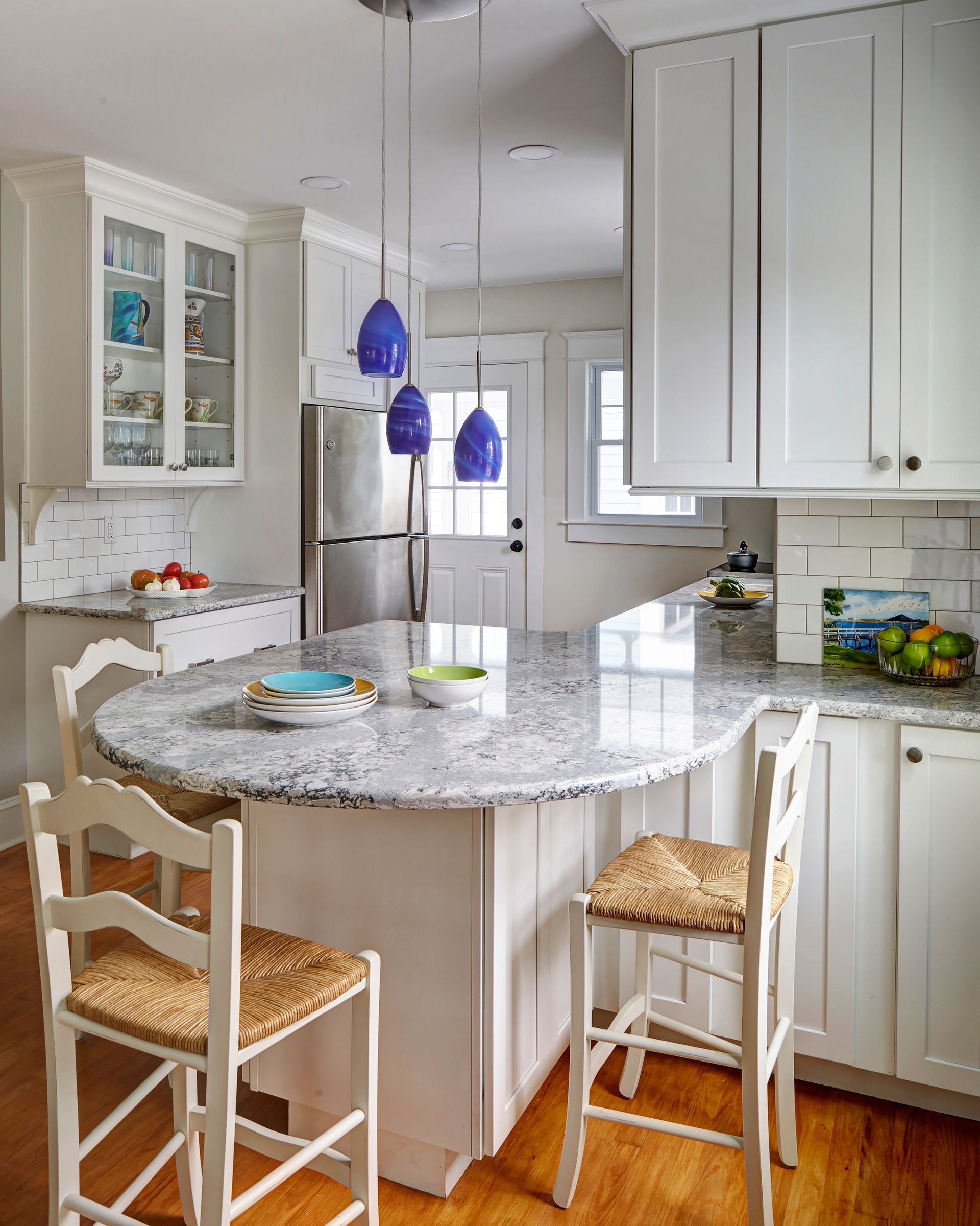 Bright white kitchen with granite island, two barstools, blue pendant lights, and stainless steel refrigerator