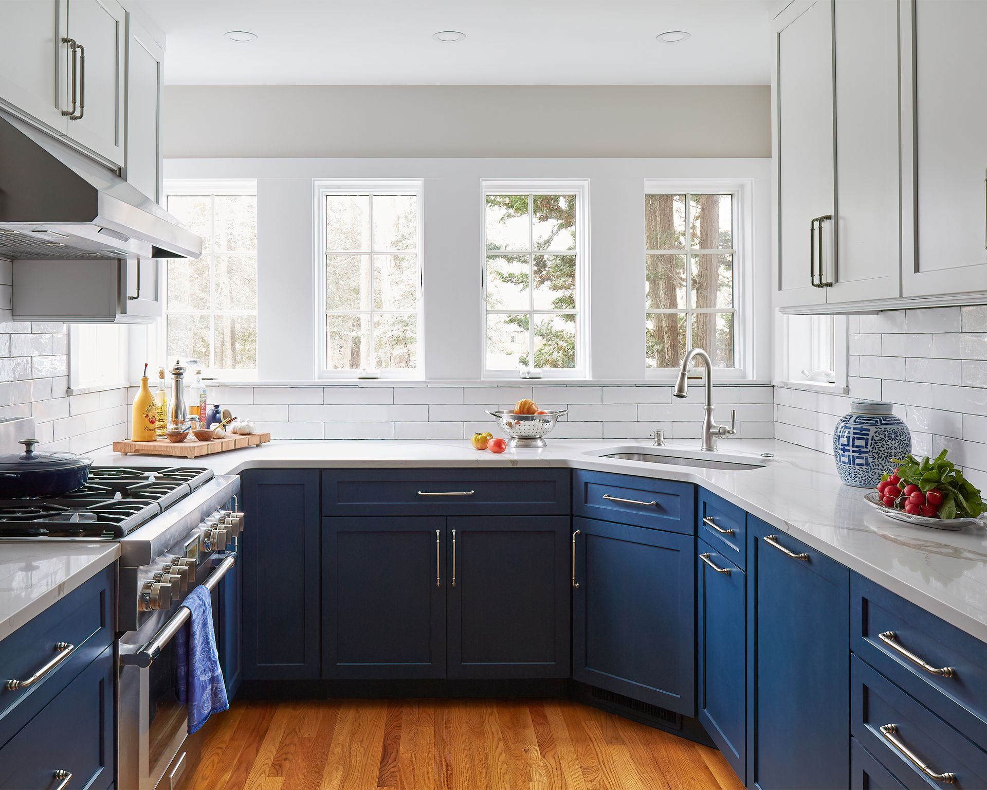 Bright kitchen with navy and white cabinets, marble counters, and large windows over a hardwood floor.