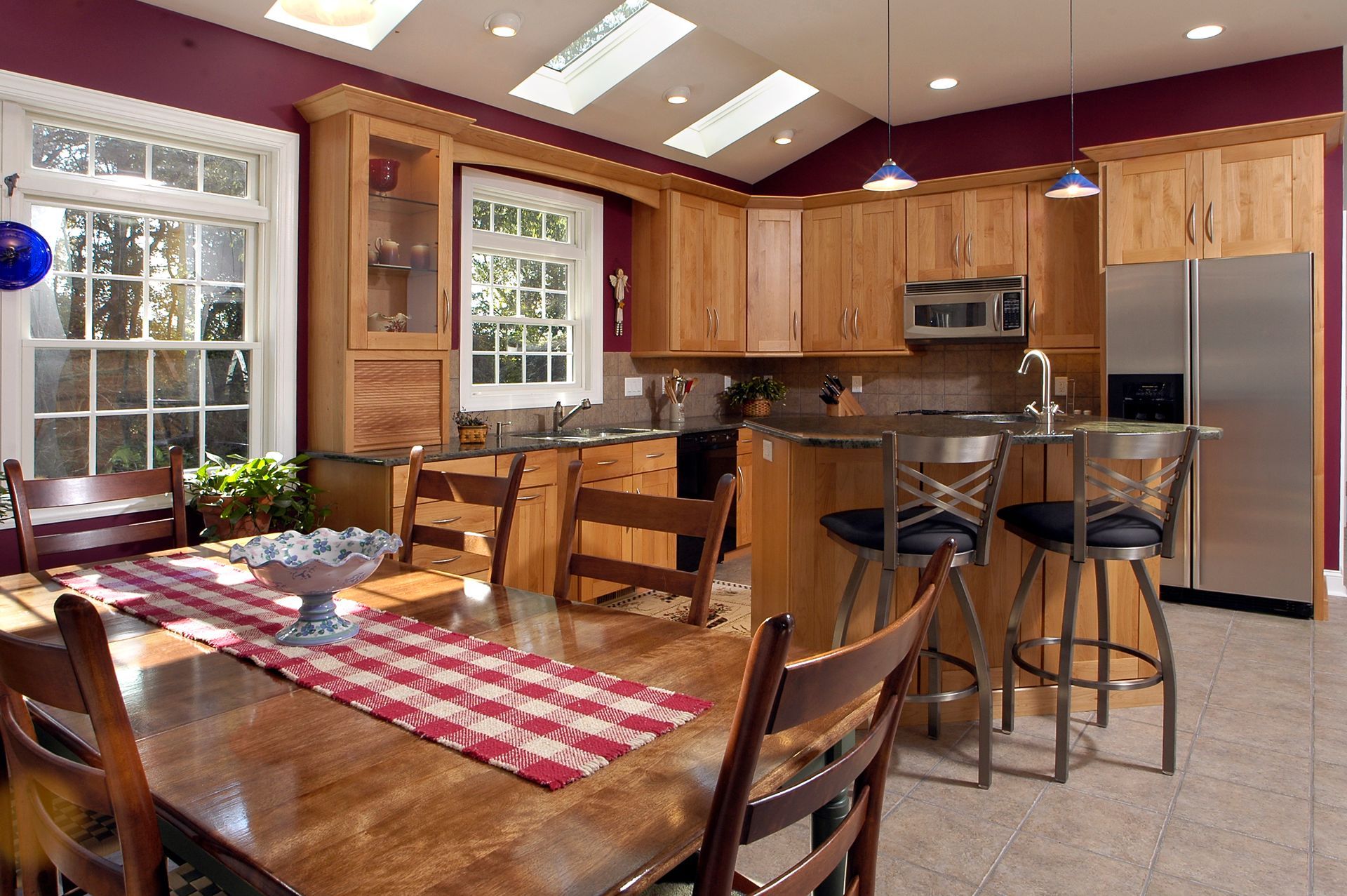 Bright kitchen with wood cabinets, island stools, dining table, and large windows with red plaid runner.