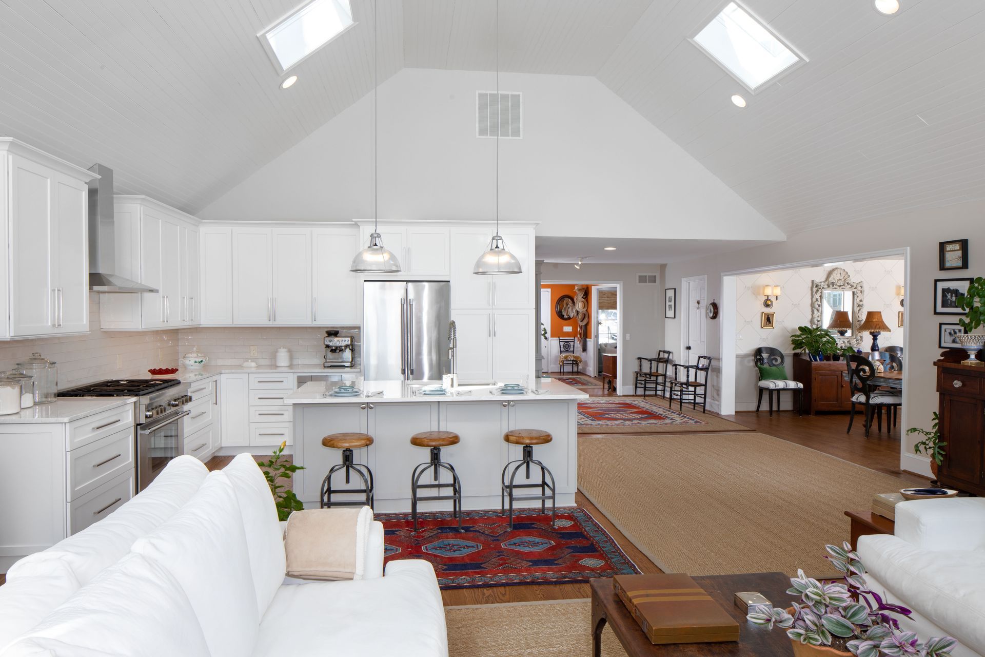 Bright, open living room and kitchen with white cabinets, skylights, island stools, and red patterned rugs.