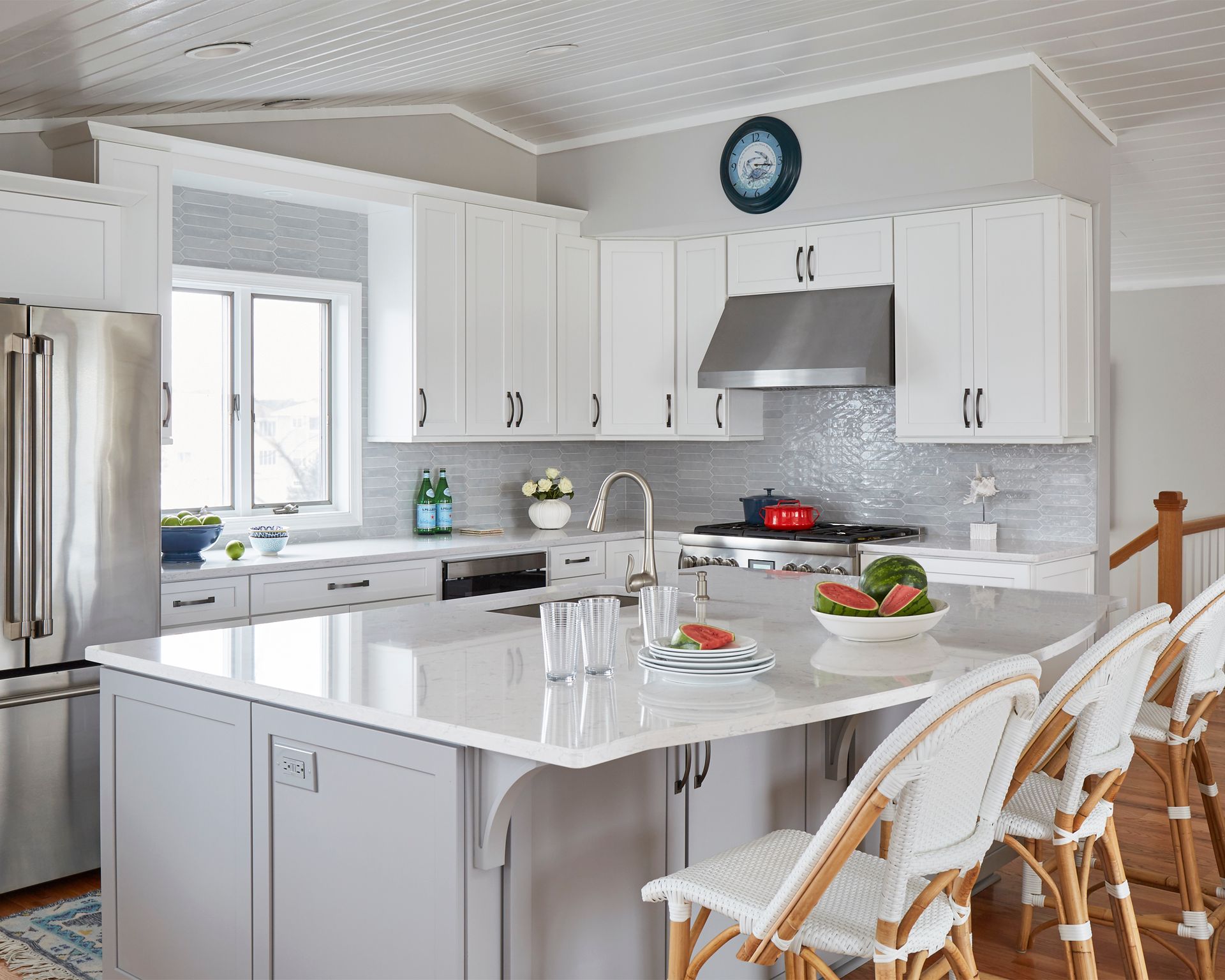 Bright white kitchen with island, stainless range hood, and wicker bar stools by a window