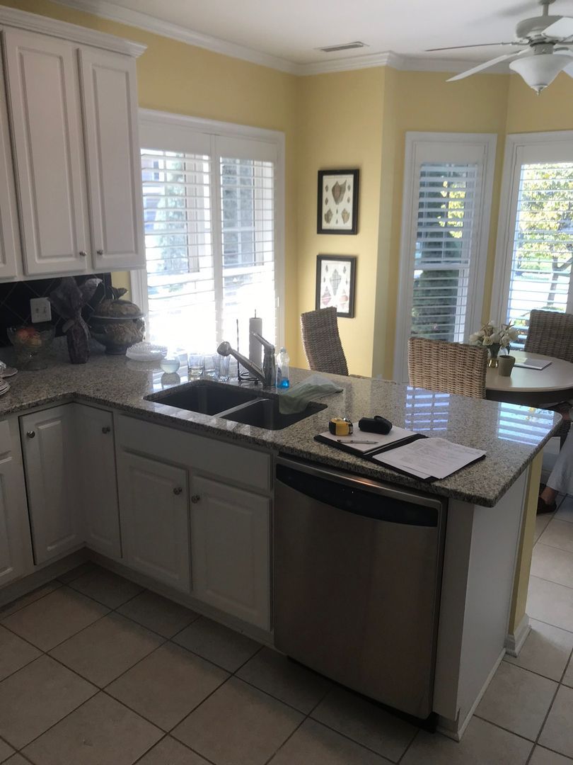 Bright kitchen with gray cabinets, granite island sink, tiled floor, and sunlit windows with wicker chairs.
