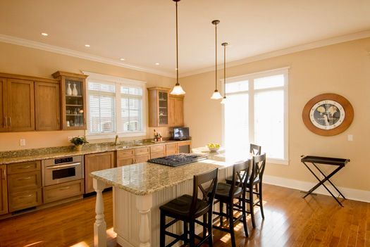 Bright kitchen with island, wood cabinets, granite counters, and dining stools by large windows.