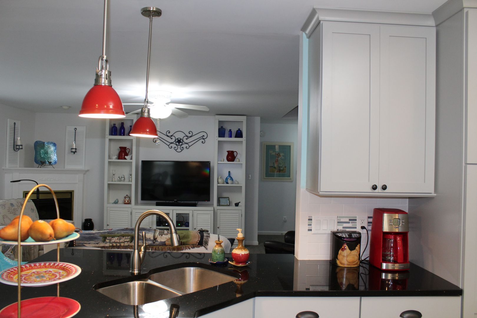 Modern kitchen with red pendant lights, black countertops, white cabinets, and a TV visible in the background