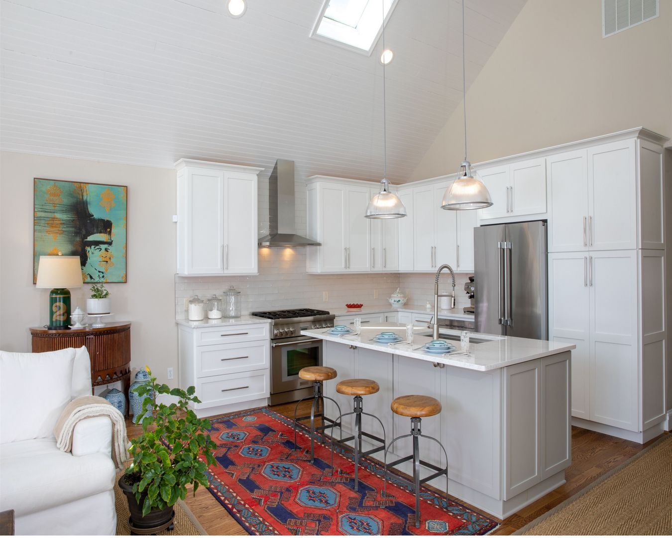 Bright open kitchen and living area with white cabinets, island stools, red rug, and vaulted ceiling