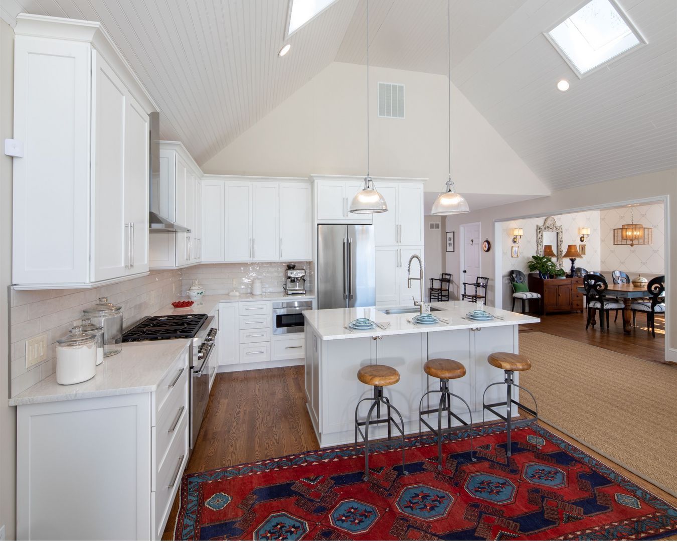 Bright modern kitchen with white cabinets, island stools, red patterned rug, and vaulted ceiling with skylights
