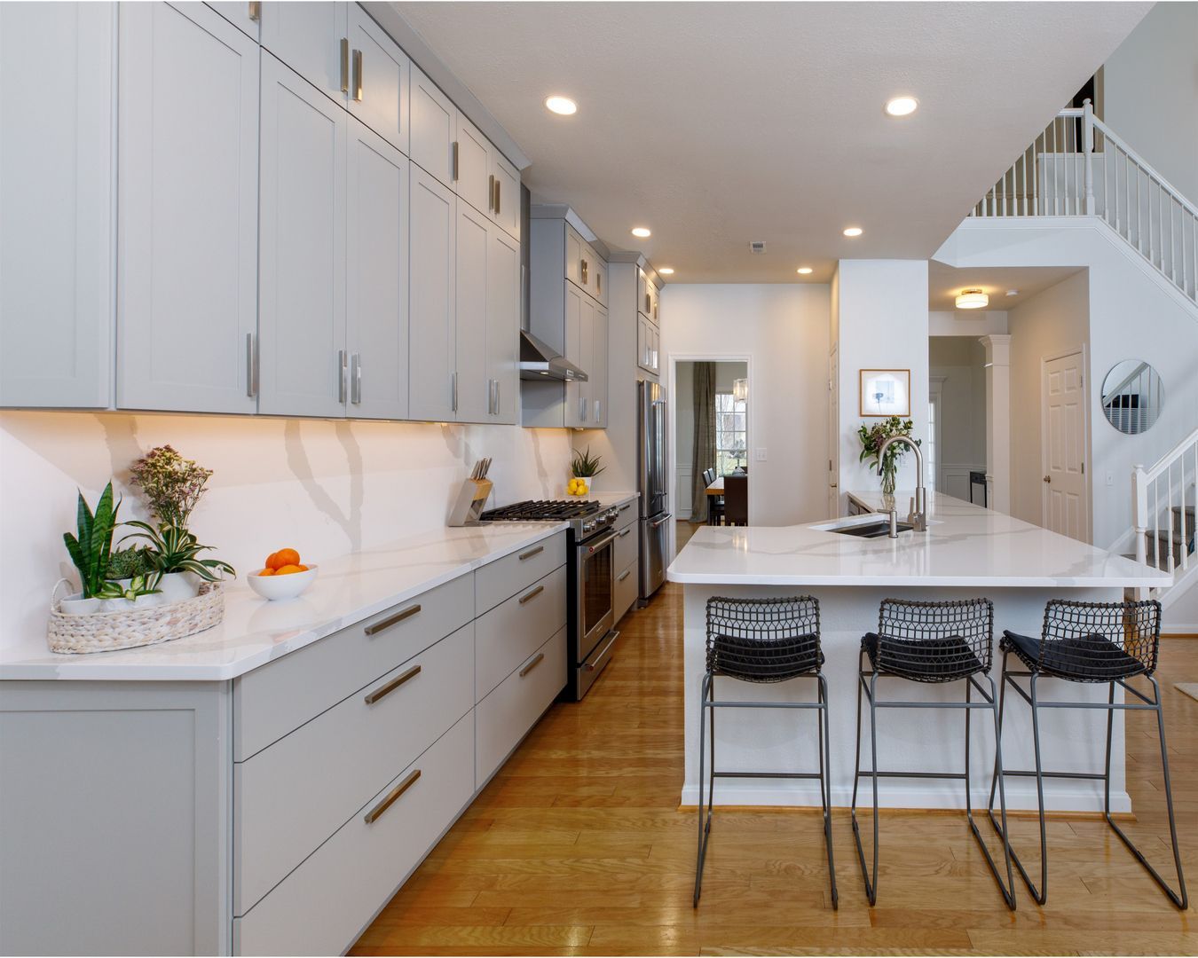 Modern bright kitchen with gray cabinets, white island, wood floors, and staircase in the background