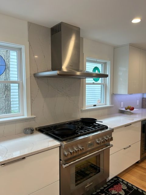 Modern kitchen with stainless steel range hood, gas stove, marble-look counters, and two windows.