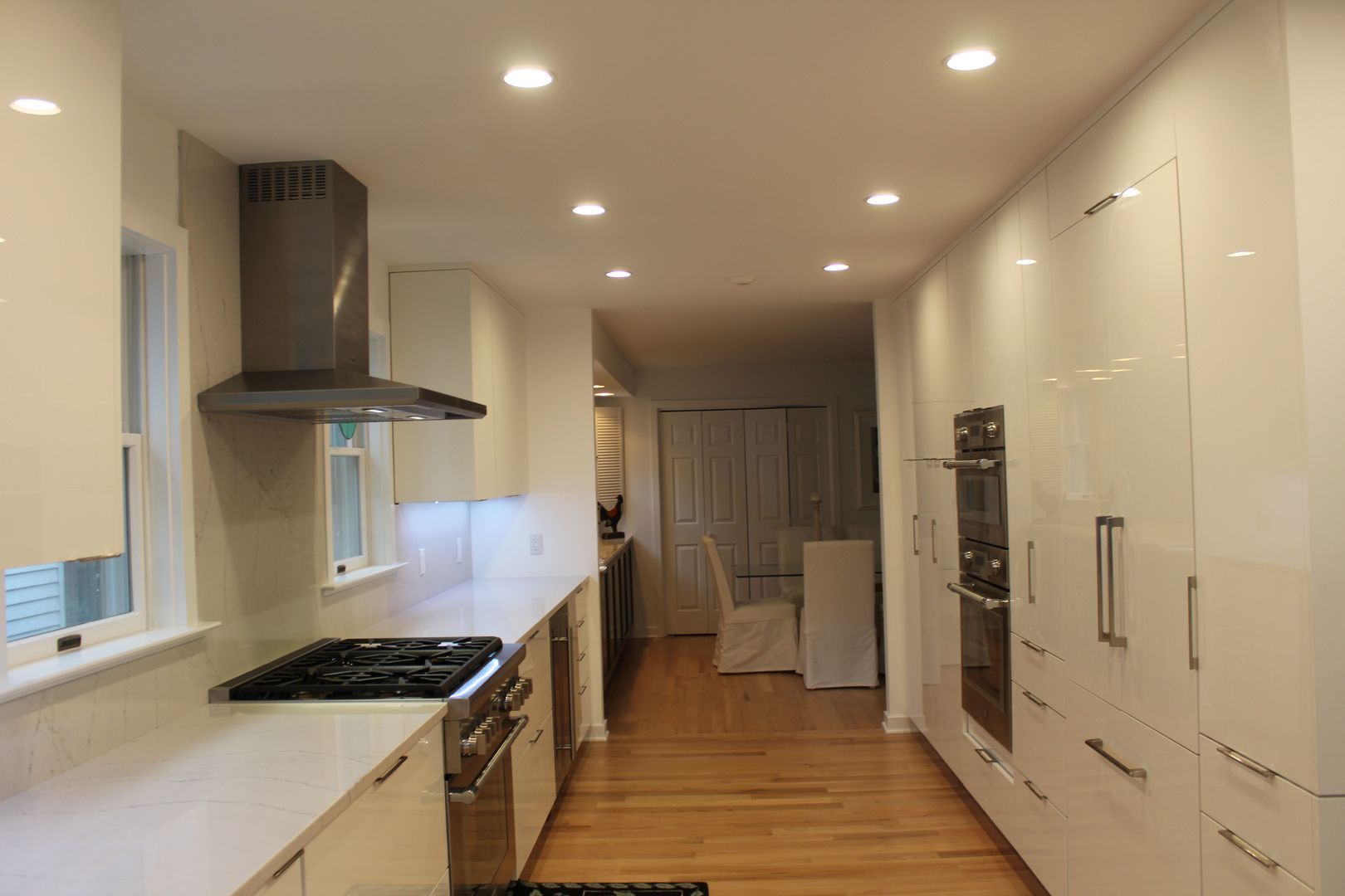 Modern white kitchen with long counters, stainless appliances, wood floor, and recessed ceiling lights