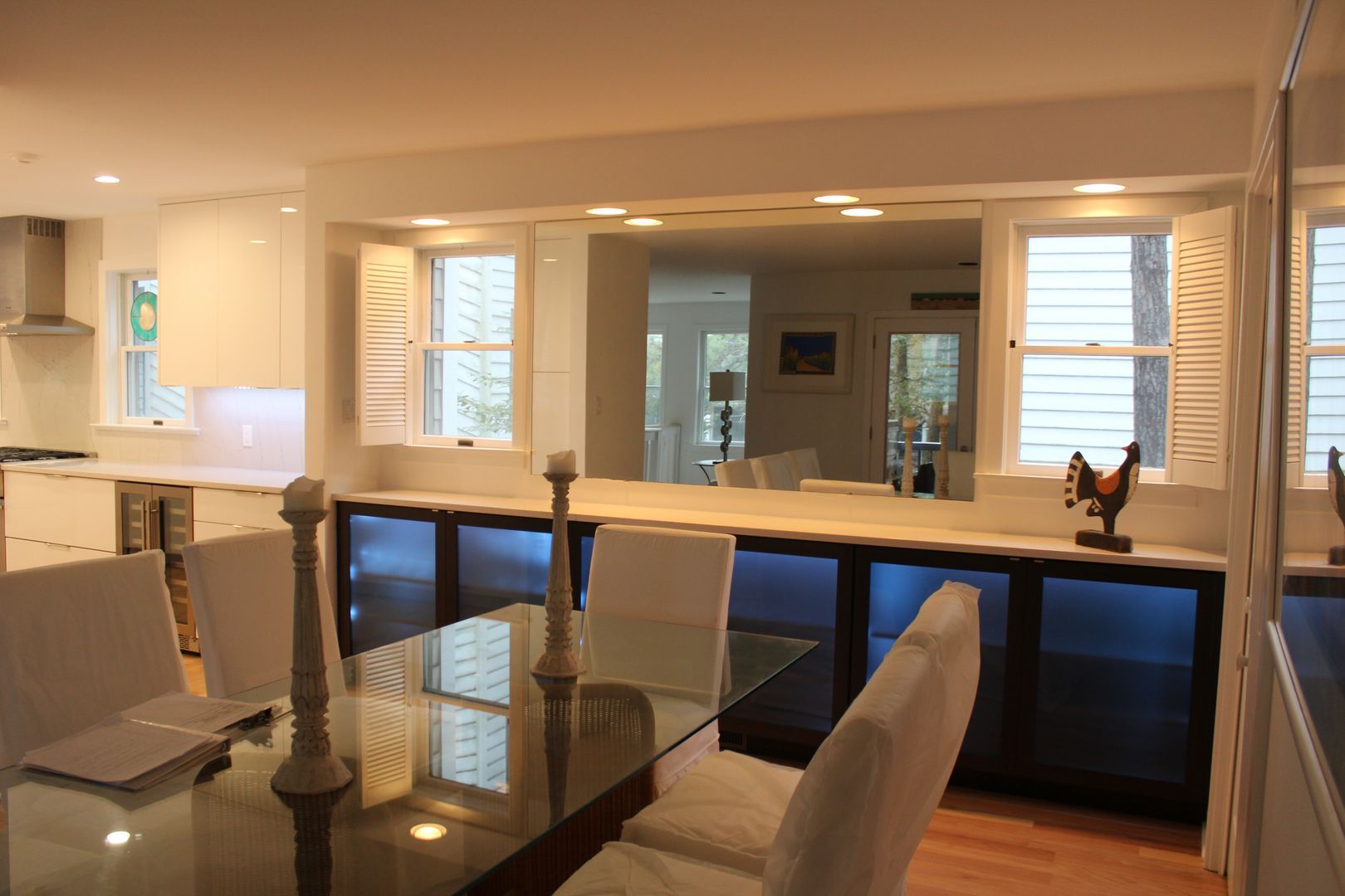 Modern kitchen and dining area with glass table, chairs, and large pass-through windows to a lit living room