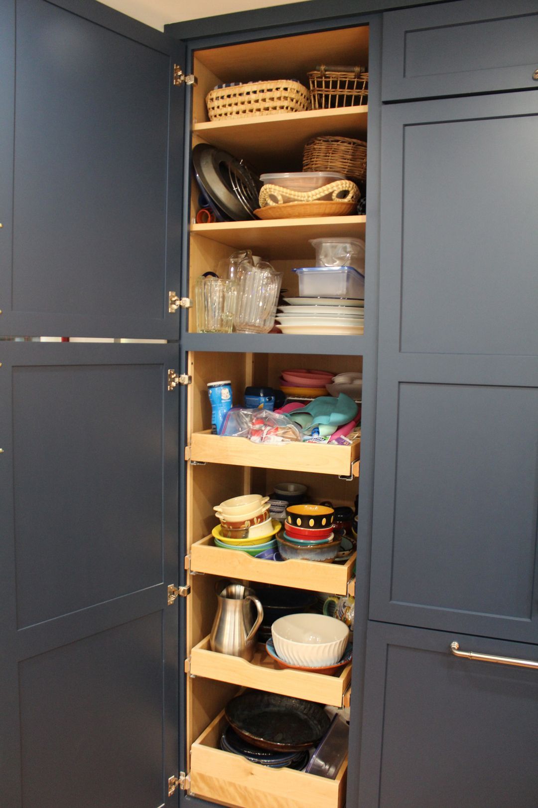 Open kitchen pantry with shelves of stacked dishes, glassware, and food containers inside dark cabinets