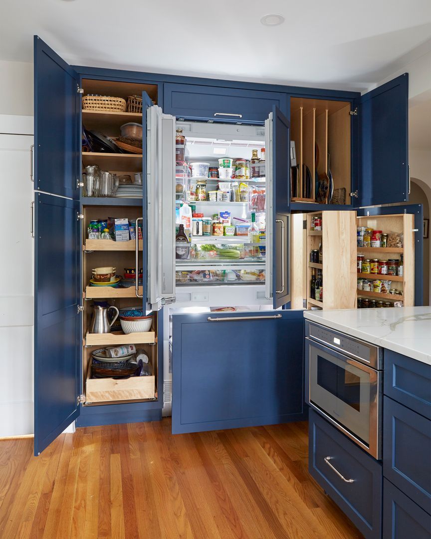 Open blue kitchen cabinets with pantry shelves, fridge, and an oven in a bright hardwood kitchen.