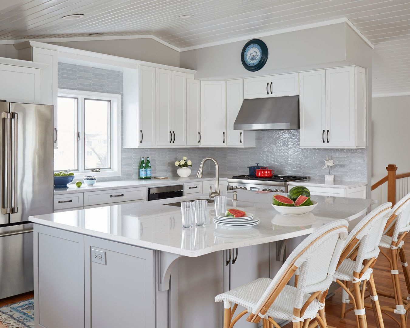 Bright white kitchen with island, stainless steel appliances, and wicker stools by a window