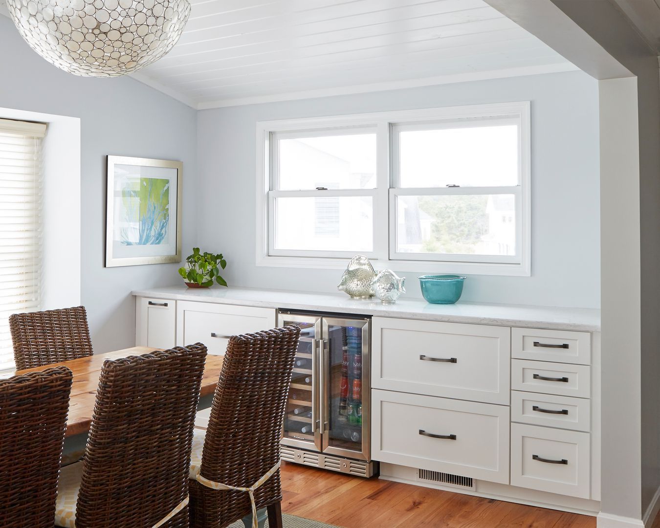 Bright dining room with woven chairs, white sideboard, blue decor, and a large window