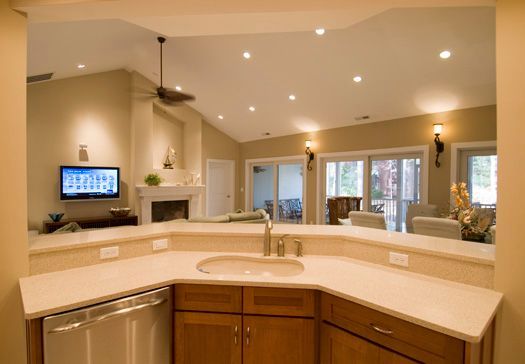Bright kitchen with curved island, beige countertops, wood cabinets, and recessed lighting, viewed from behind a sink.