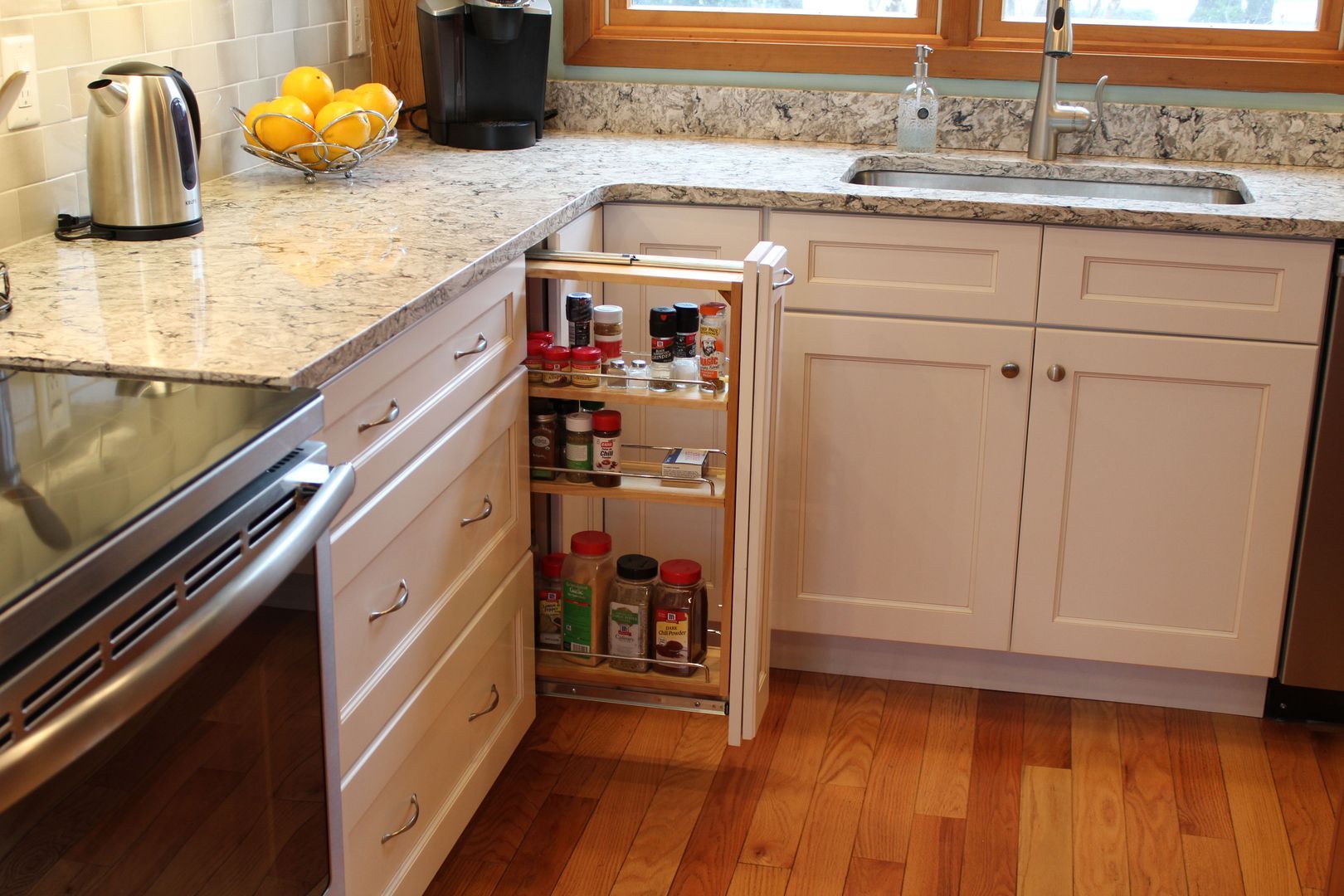 Kitchen with open spice cabinet under the counter beside a stainless stove and sink, with fruit on the counter.