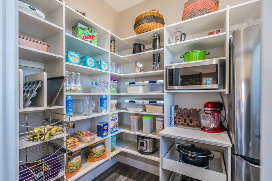 Pantry closet with white shelves, stocked food, baskets, and kitchen appliances, next to a stainless steel refrigerator