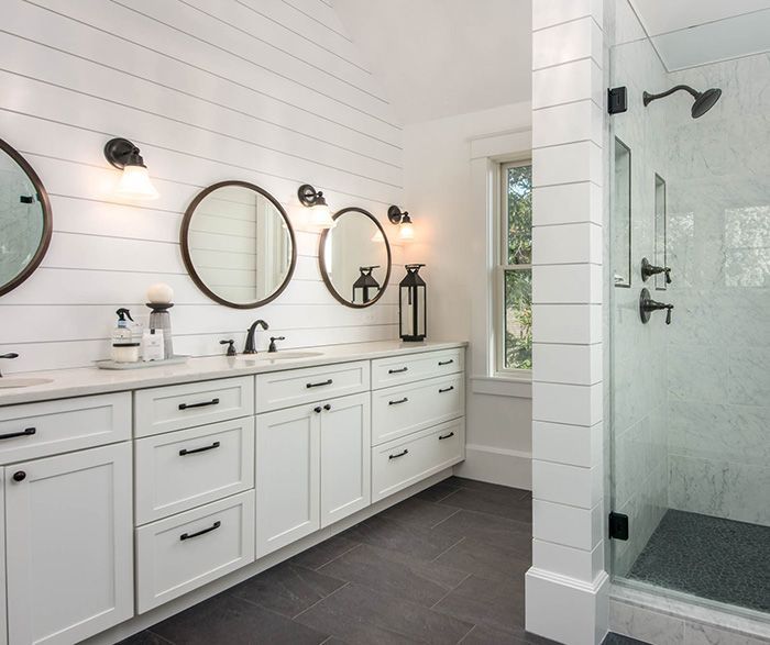 Bright white bathroom with double vanity, round mirrors, wall sconces, and glass shower