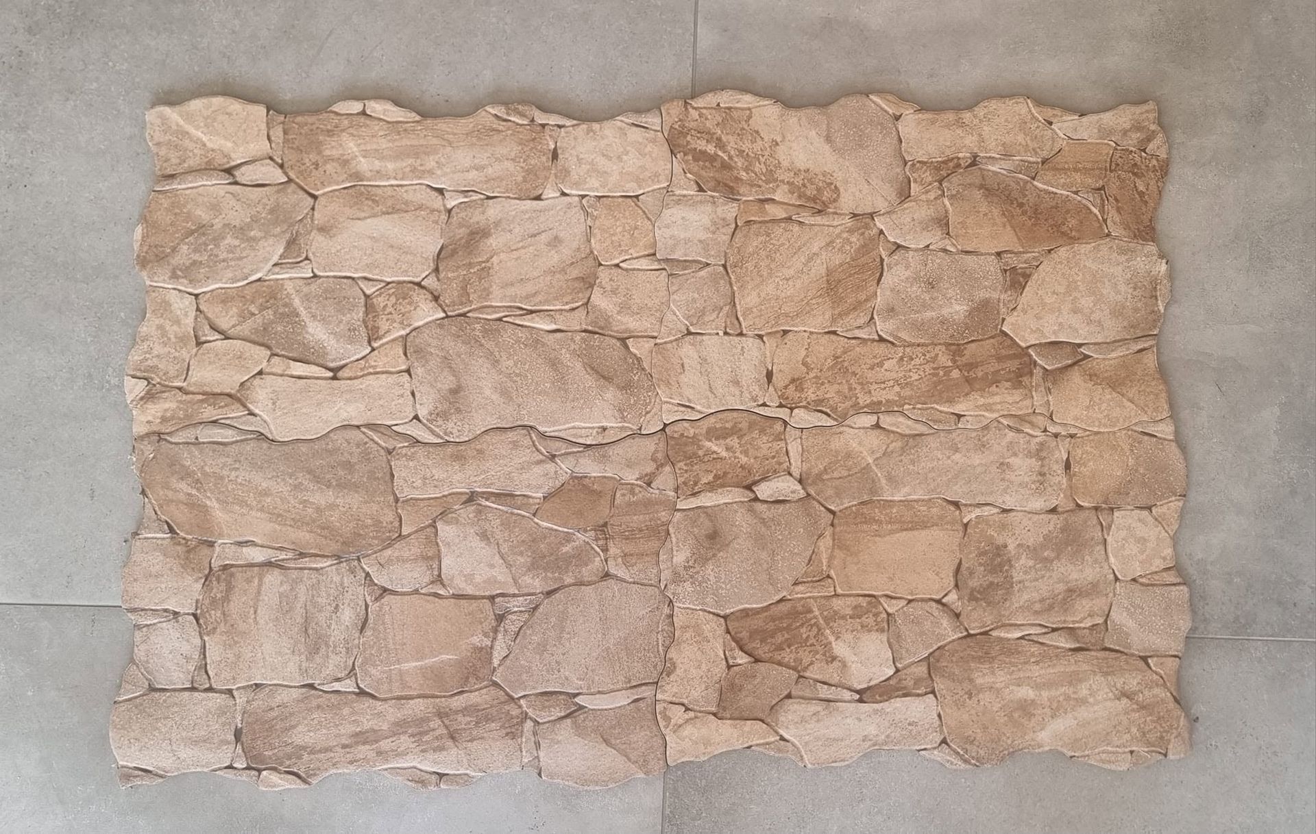 Pannello murale in pietra color sabbia con pietre irregolari su sfondo grigio.