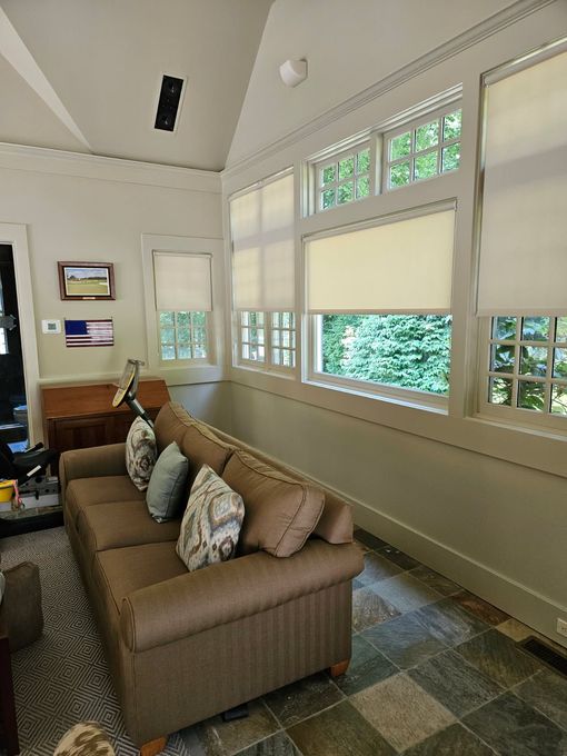 A brown couch with decorative pillows sits in a sunroom with large windows, light walls, and stone tile flooring.