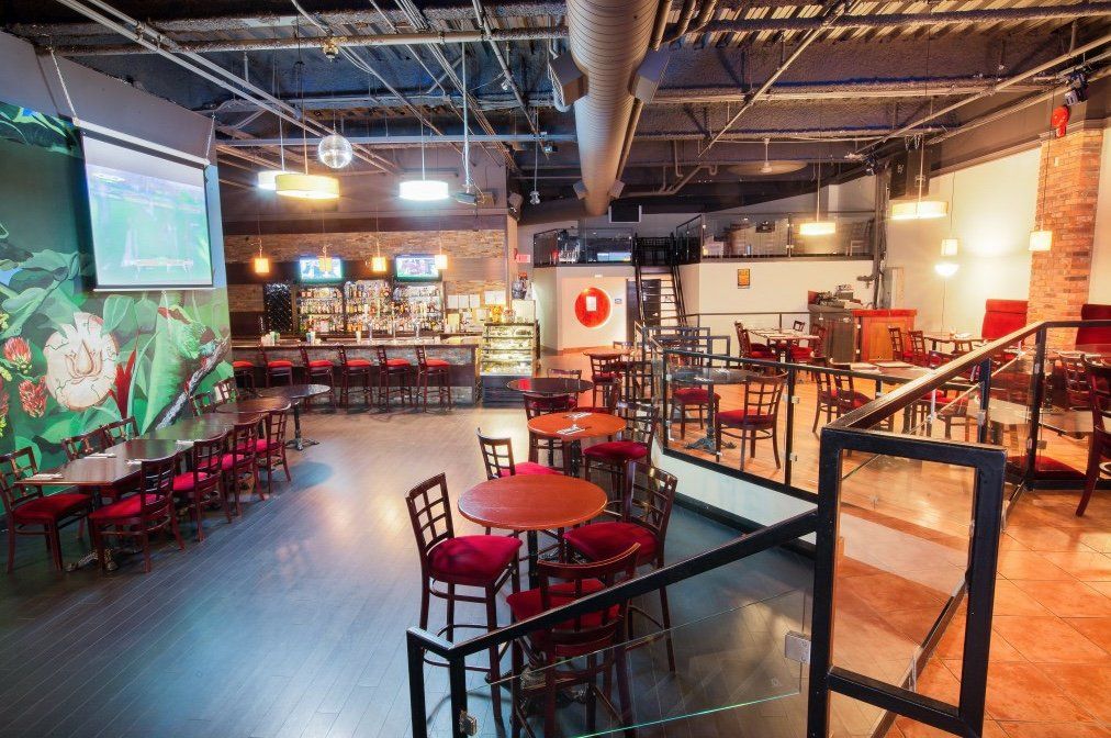 Interior of a bar/lounge with tables, chairs, bar, and a projection screen. Red chairs and tables, exposed brick walls.