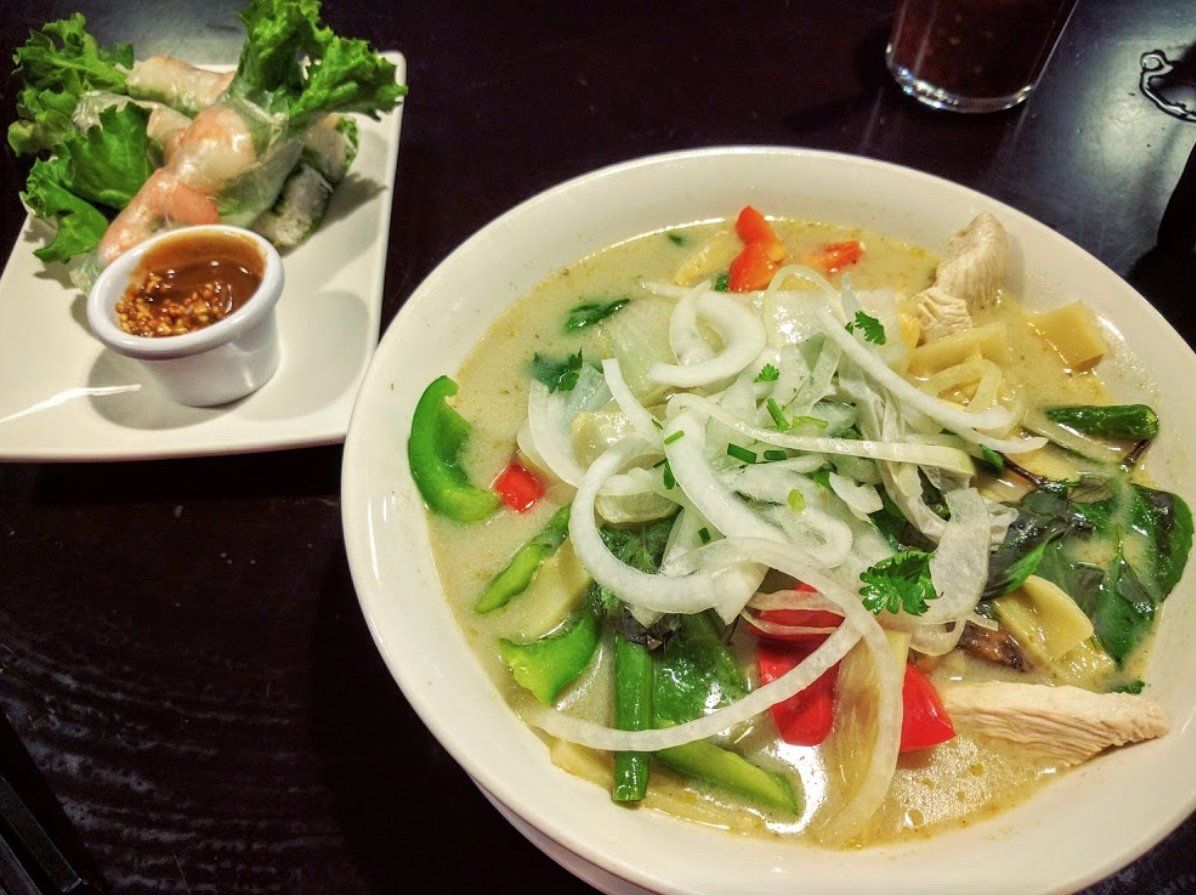 Bowl of green curry with vegetables and onion, spring rolls with dipping sauce on a white plate.