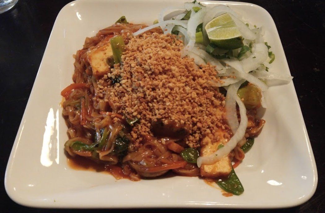 Pad Thai with peanuts, tofu, and onions on a white square plate.
