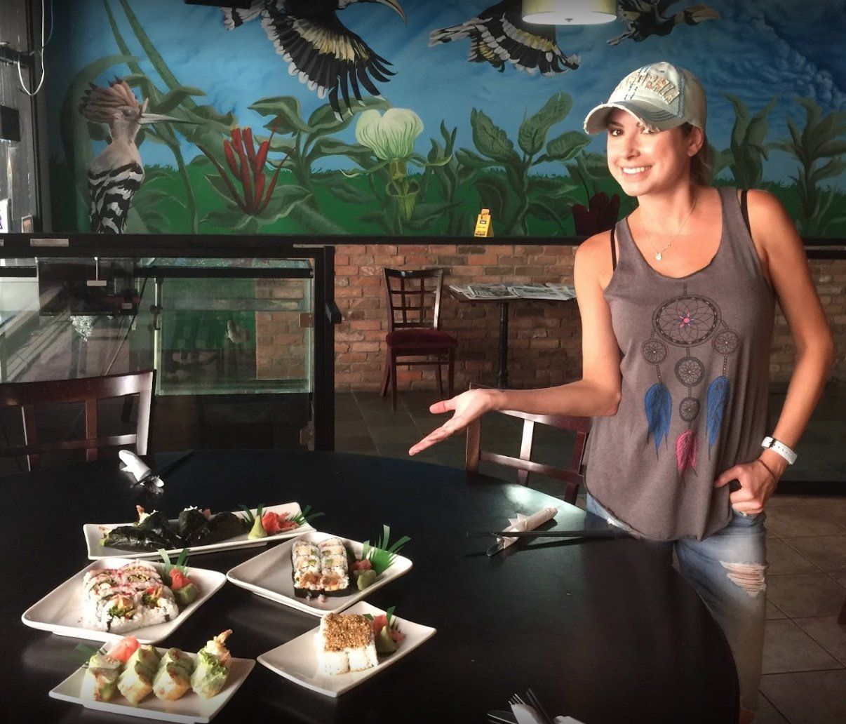 Woman pointing at sushi plates on a table in a restaurant with a mural background.