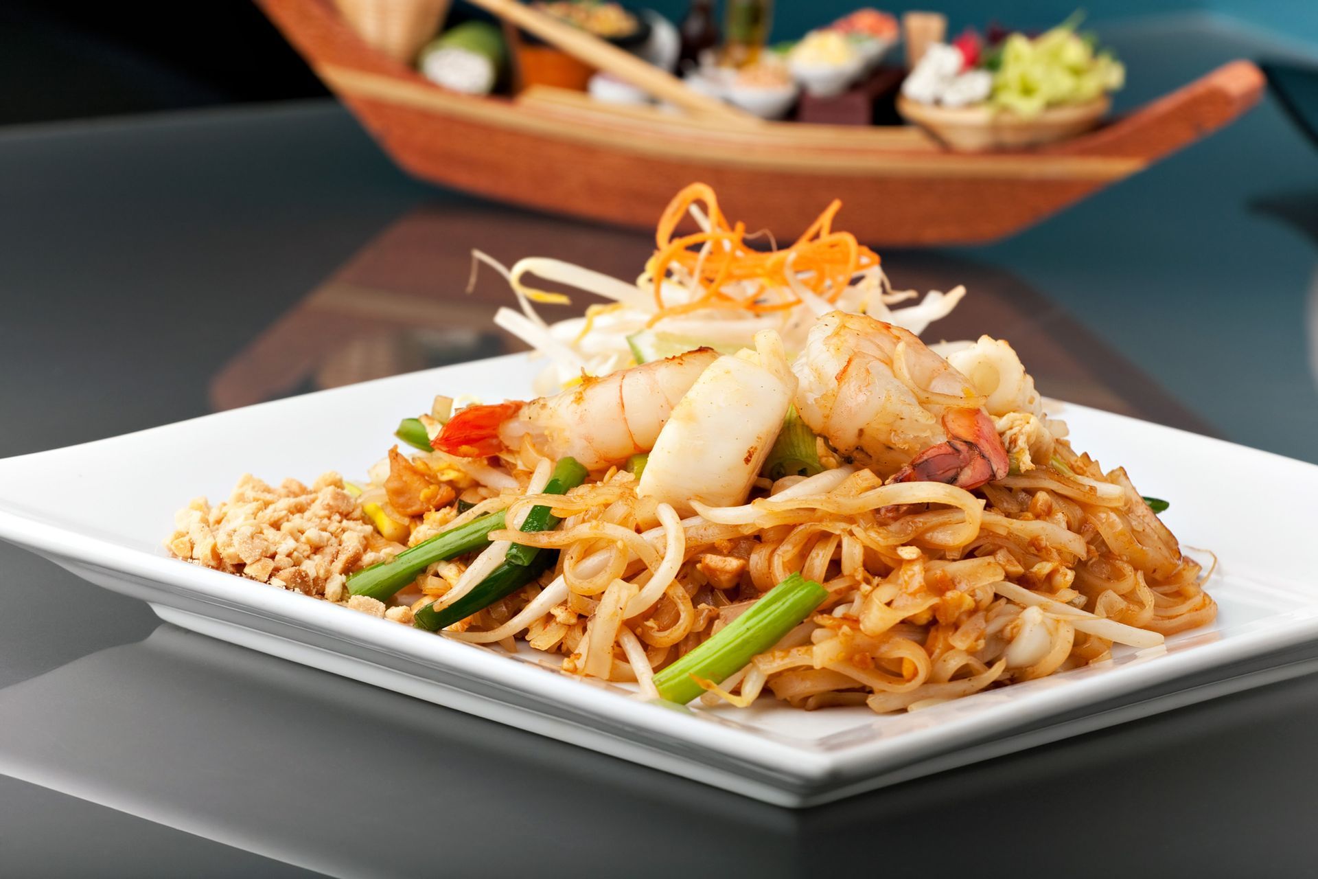 Pad Thai with shrimp on a white square plate, sushi boat in the background.