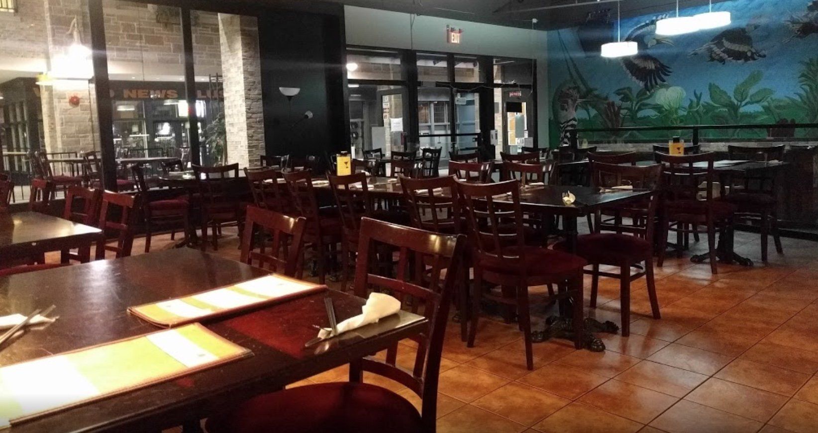 Empty restaurant interior with brown tables, chairs, and dark walls. Large windows look outside.