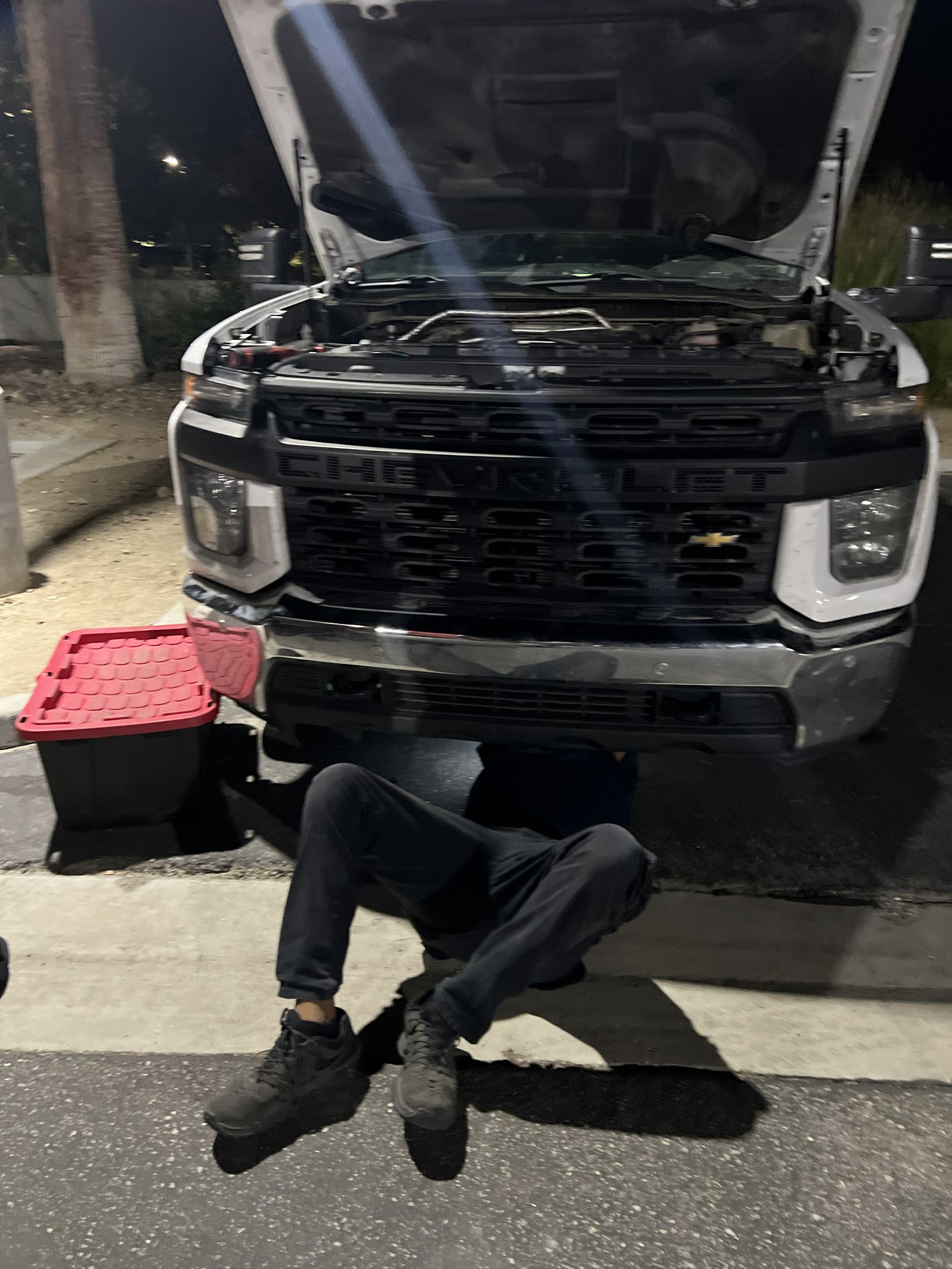 Person lying under the front of a black pickup truck in a dim garage, with a red toolbox nearby.
