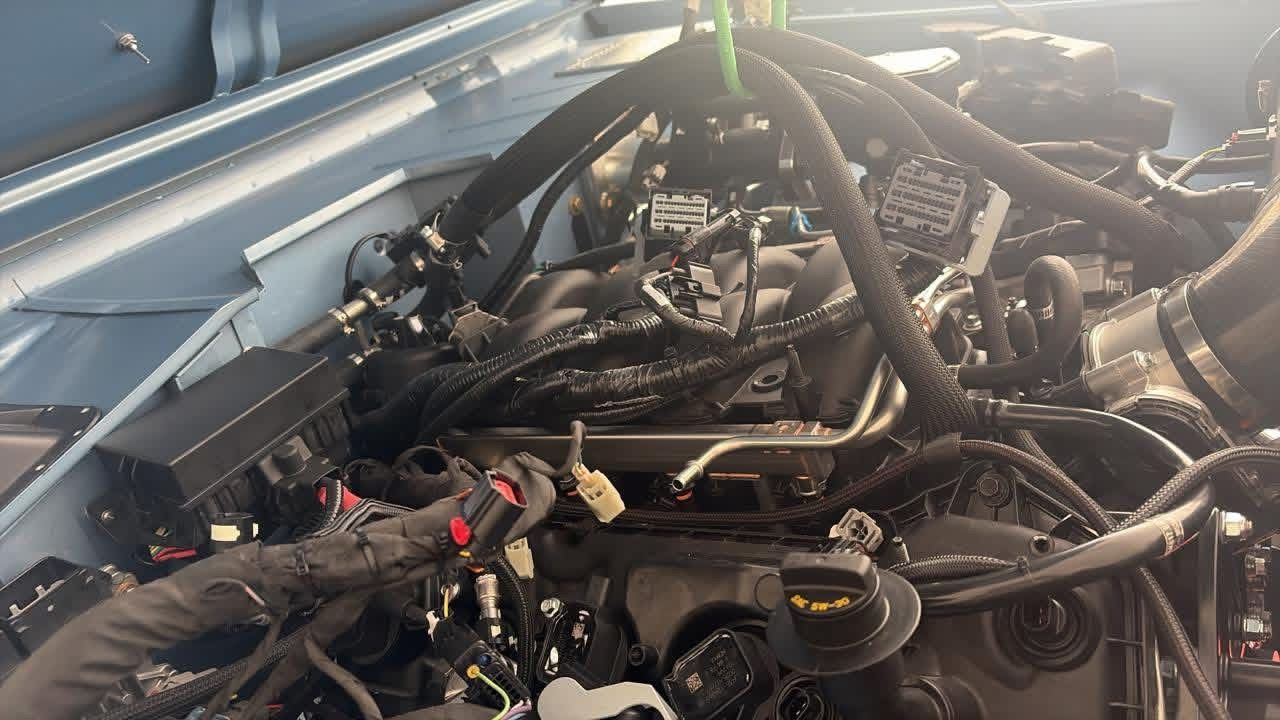 Car engine bay with exposed wiring and hoses, viewed from above under the hood.