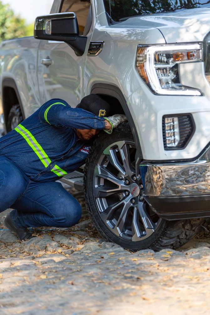 Tow truck worker in blue uniform kneels beside a silver pickup truck’s front wheel on a sandy road.