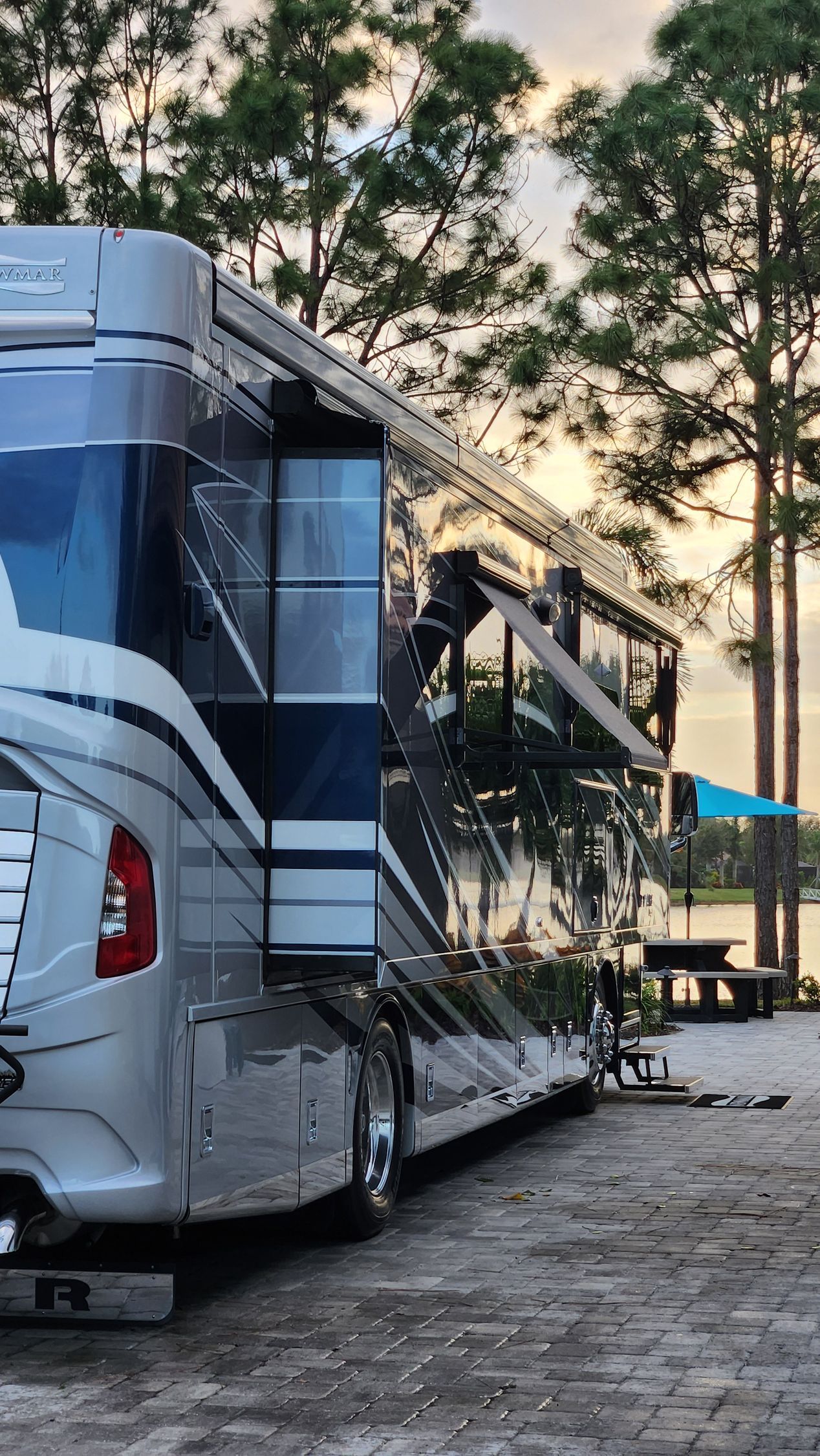 Shiny silver RV parked near water with a slide-out, under a backdrop of trees, evening sky.